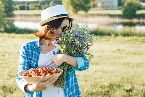 成人妇女的户外夏季肖像与草莓, 野花花束, 草帽和太阳镜。自然背景, 乡村景观, 绿色草地, 乡村风格 — 图库照片