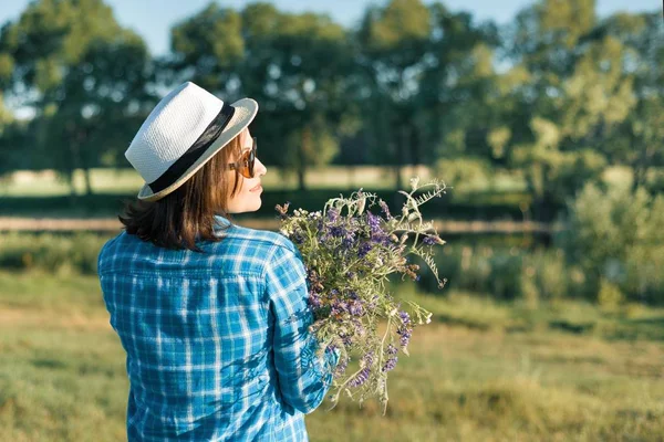 Ritratto estivo esterno di donna con bouquet di fiori di campo, cappello di paglia. Vista dal retro, sfondo naturale, paesaggio rurale, prato verde, stile country . — Foto Stock