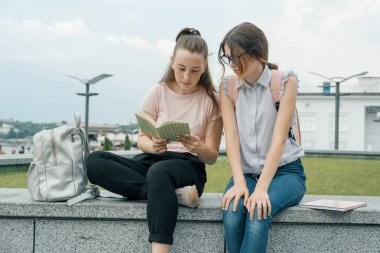Sırt çantası, kitap ile iki genç güzel kız öğrencilerin açık portre. Kızlar konuşuyor, bir kitaba bakarak, kentsel arka plan