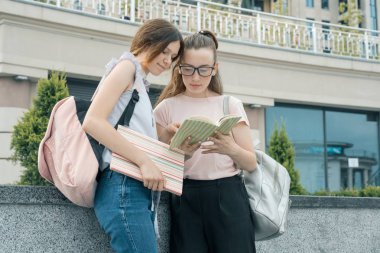 Sırt çantası, kitap ile iki genç güzel kız öğrencilerin açık portre. Kızlar konuşuyor, bir kitaba bakarak, kentsel arka plan