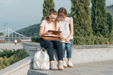 Sırt çantası, kitap ile iki genç güzel kız öğrencilerin açık portre. Kızlar konuşuyor, bir kitaba bakarak, kentsel arka plan