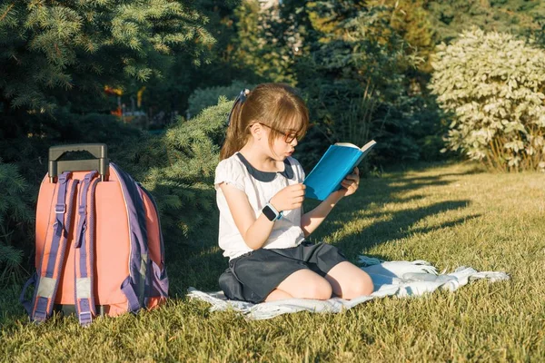 Маленька дівчинка читає книгу з шкільним рюкзаком, що сидить на траві в парку — стокове фото