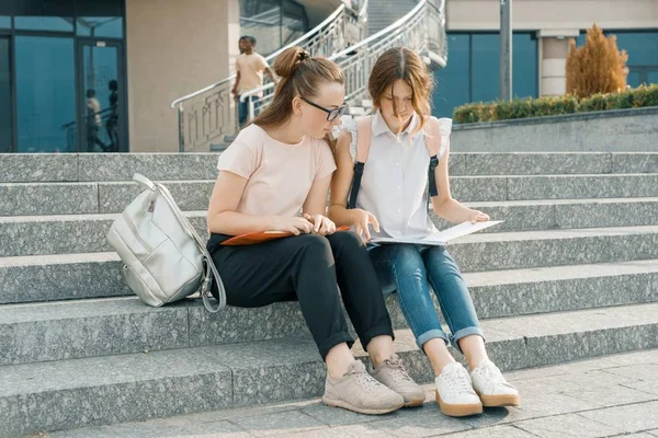 Retrato ao ar livre de dois jovens estudantes meninas bonitas com mochilas, livros. Meninas sentadas nos degraus, falando, olhando para um livro — Fotografia de Stock