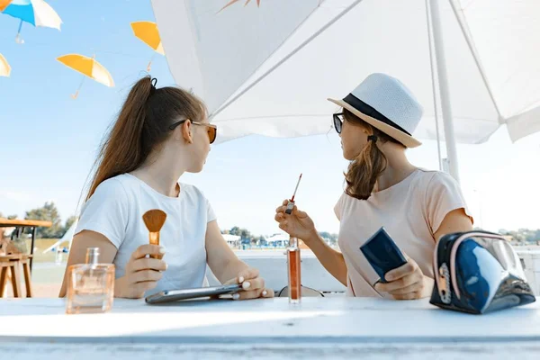 Las jóvenes adolescentes que se divierten en un café al aire libre haciendo maquillaje usan brillo de labios, pincel, perfume. Café de verano calle, área de recreación, fondo del parque de la ciudad, estilo de vida juvenil — Foto de Stock