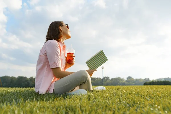 Volwassen vrouw rusten in het Park, zittend op het gras met boek en zomer verfrissing drinken, gouden uur — Stockfoto