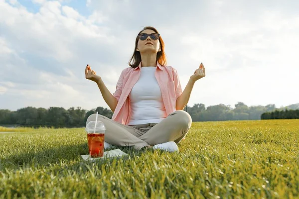 Зрелая женщина практикует йогу, сидя на траве. Женщина в парке с летним напитком, книгой, отдыхом и медитацией, золотой час — стоковое фото