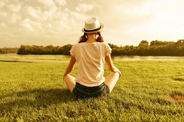 年轻的少女坐在河边的公园里, 后眼景, 女孩看着水, 练习瑜伽, 冥想 — 图库照片