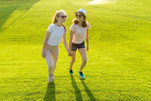 Dvě dívky se drží na zelené trávě v parku za horkého letního dne ve zlaté hodině. Přátelství mezi dospívající — Stock fotografie