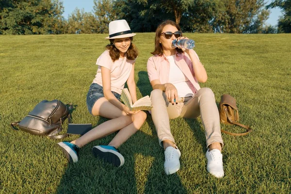 Мать и дочь, улыбающаяся мать и девочка-подросток гуляют в летнем парке, пьют напитки, отдыхают на траве, золотой час — стоковое фото