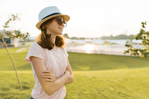 Outdoor portrait of young girl in sunglasses with crossed arms. Background of green grass, recreation area, summer sunset. Copy space, golden hour — Stock Photo, Image
