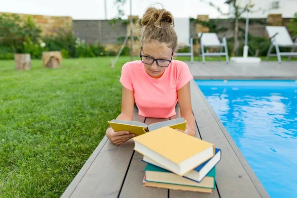 Молодая девушка в очках у бассейна с грудой книг, читающая книгу. Образование, лето, знания — стоковое фото