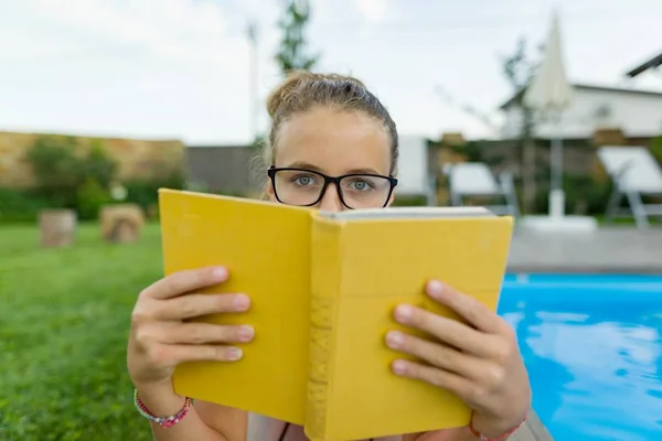 Девочка-подросток в очках читает книгу, плавательный бассейн на заднем плане, газон рядом с домом. Школа, образование, знания, образование — стоковое фото