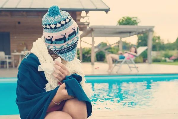 La chica cogió un resfriado, vistiendo un cálido sombrero de punto, ropa con un pañuelo, mira a su amiga descansando en un sillón junto a la piscina — Foto de Stock