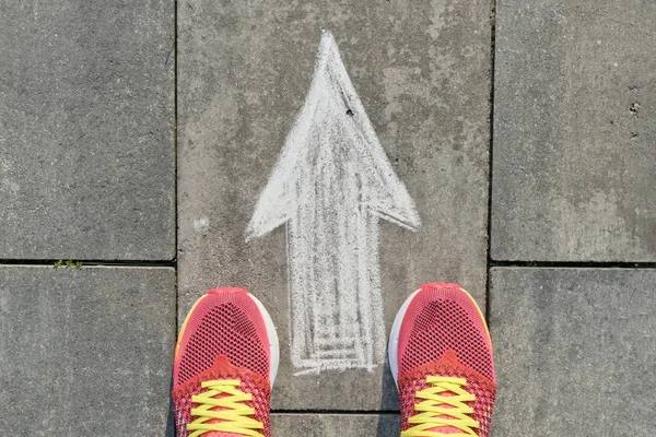 Βέλος σύμβολο ζωγραφισμένο σε γκρίζο πεζοδρόμιο με τα πόδια των γυναικών σε αθλητικά παπούτσια, κορυφή θέα — Φωτογραφία Αρχείου