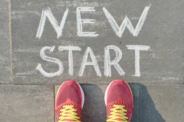 Palabra nuevo comienzo escrito en pavimento gris con piernas de mujer en zapatillas de deporte, vista desde arriba — Foto de Stock