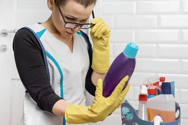 Доросла жінка робить чистку за допомогою миючих засобів. Жінка в окулярах, професійна форма у ванній, туалетній кімнаті, читання інструкцій по прибиранню пляшки — стокове фото