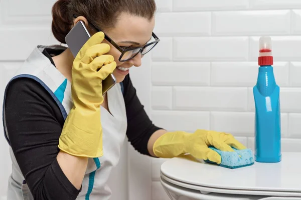 Взрослая женщина занимается уборкой моющими средствами. Женщина в очках, профессиональная униформа в ванной, туалеты, женщина смеется и разговаривает по телефону — стоковое фото