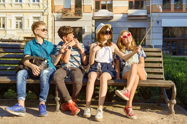 Feliz 4 amigos adolescentes o estudiantes de secundaria se están divirtiendo, hablando, leyendo el teléfono, haciendo foto selfie en la ciudad en el banco. Concepto de amistad y personas — Foto de Stock