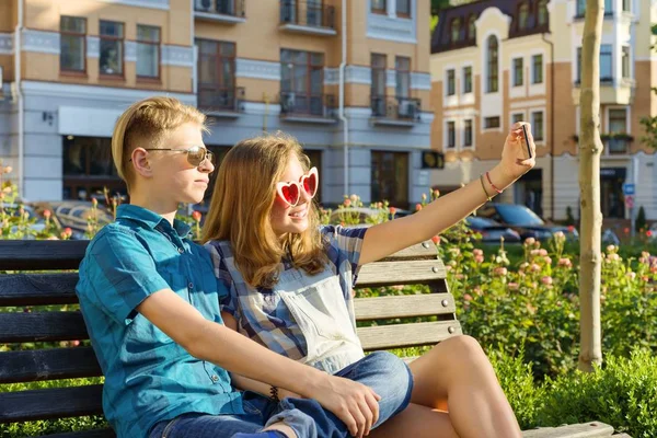 Tizenéves barátok lány és fiú ül a padon a városban, beszél, keresi a telefont, ezzel selfie fotó — Stock Fotó