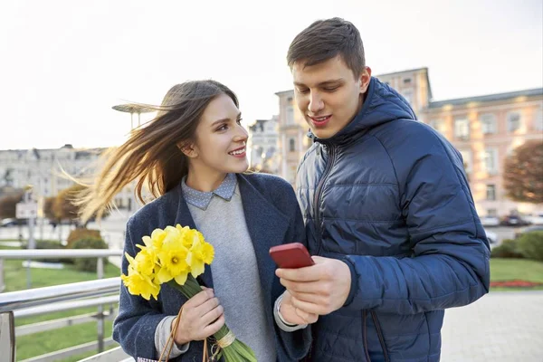 Kültéri portréja gyönyörű romantikus pár, fiatal férfi és a nő a csokor sárga virágok Nárciszok és keres a smartphone, tavaszi város háttér — Stock Fotó