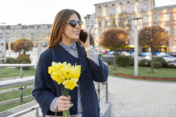 Beautiful girl with bouquet of yellow spring flowers, young woman talking on the phone, urban style background. — Stock Photo, Image