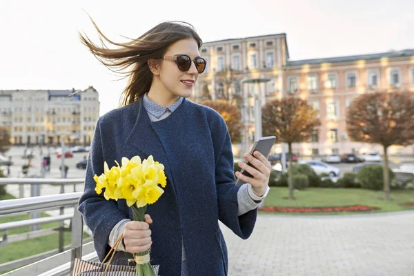 Beautiful girl with bouquet of yellow spring flowers, young woman reading text with smartphone, background city architecture, golden hour — Stock Photo, Image