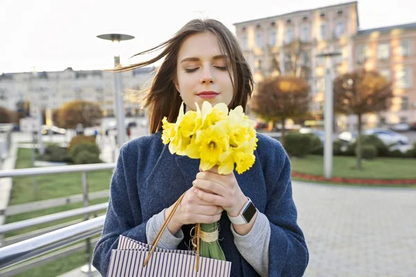 Портрет молодой женщины с букетом желтых весенних цветов нарциссов. Красивая девушка в городе наслаждается цветами, глаза закрыты . — стоковое фото