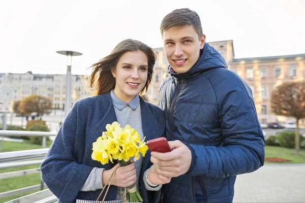 Εξωτερική πορτρέτο του όμορφο ρομαντικό ζευγάρι, νέος άνδρας και γυναίκα με μπουκέτο κίτρινα άνθη των νάρκισσων και κοιτάζοντας στο smartphone, άνοιξη φόντο της πόλης — Φωτογραφία Αρχείου