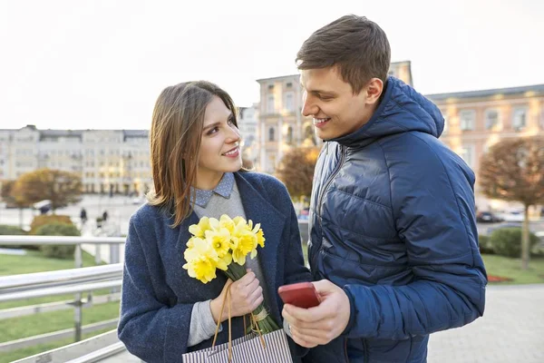 Utomhus porträtt av vackra romantiska par, ung man och kvinna med bukett gula blommor av påskliljor och titta i smartphone, Spring City bakgrund — Stockfoto