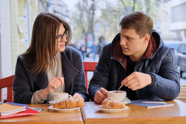 Jóvenes amigos y amigas estudiantes sentados en la cafetería al aire libre, hablando, tomando café, té, comiendo croissants. En los libros de texto de mesa, cuadernos, fondo de la ciudad . — Foto de Stock