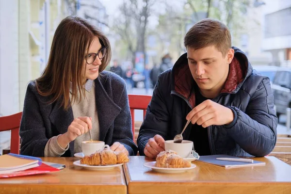 Jóvenes amigos y amigas estudiantes sentados en la cafetería al aire libre, hablando, tomando café, té, comiendo croissants. En los libros de texto de mesa, cuadernos, fondo de la ciudad . — Foto de Stock