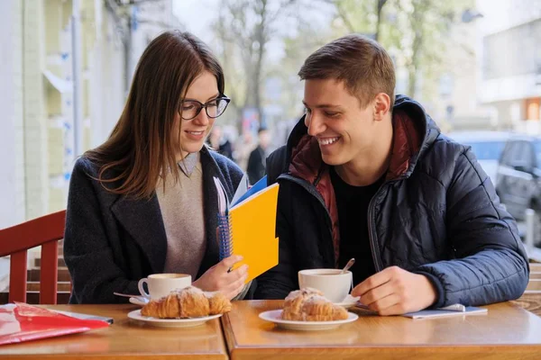 Young male and female friends students sitting in outdoor cafe, talking, drinking coffee, tea, eating croissants. On table textbooks, notebooks, city background. — 스톡 사진