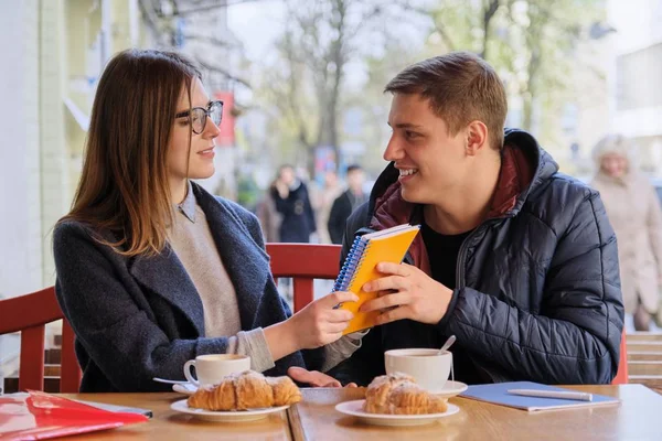 Young couple of students study in outdoor cafe, drink coffee tea, eat croissants, background is spring city street — 스톡 사진