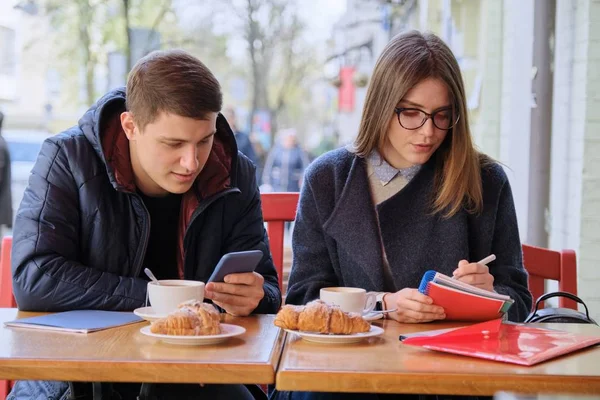 Young couple of students study in outdoor cafe, drink coffee tea, eat croissants, background is spring city street — 스톡 사진