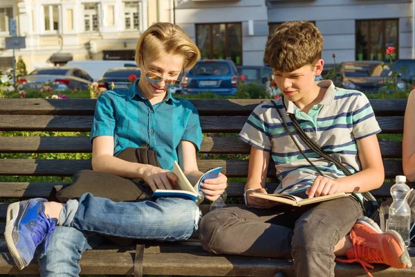Улыбаясь, двое школьников читают книги, сидя на скамейке в городе — стоковое фото