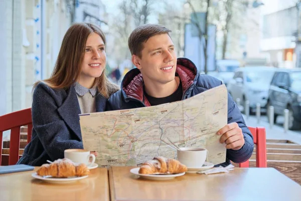 Νέοι τουρίστες άνδρας και γυναίκα ανάγνωση χάρτη της πόλης σε υπαίθριο καφέ. Ζευγάρι πίνοντας τσάι καφέ και τρώγοντας κρουασάν, άνοιξη φόντο της πόλης — Φωτογραφία Αρχείου