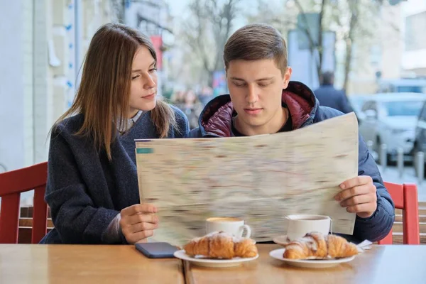 Young tourists man and woman reading map of city in outdoor cafe. Couple drinking coffee tea and eating croissants, spring city background — 스톡 사진