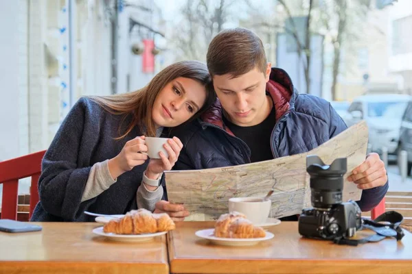 Junges schönes Touristenpaar, das sich im Café im Freien ausruht, Stadtplan liest, Kaffee mit Croissants trinkt. — Stockfoto