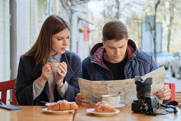 Young tourists man and woman reading map of city in outdoor cafe. Couple drinking coffee tea and eating croissants, spring city background — 스톡 사진