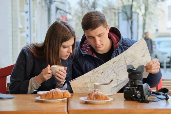 Young beautiful couple of tourists resting in outdoor cafe, reading city map, drinking coffee with croissants. — 스톡 사진