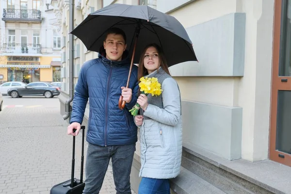 Αγαπούν το ζευγάρι των τουριστών με βαλίτσα κάτω από μια ομπρέλα. Νέος χαμογελαστός άνθρωπος και γυναίκα, κορίτσι με ανθοδέσμη, άνοιξη φόντο της πόλης. — Φωτογραφία Αρχείου