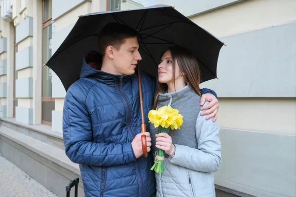 一对爱在伞下带着手提箱的游客。年轻的微笑男人和女人，女孩与花束，春天的城市背景. — 图库照片