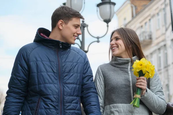 Couple in love in city. Young happy man and woman in spring city talking, walking, with bouquet of yellow flowers daffodils — Stock Photo, Image