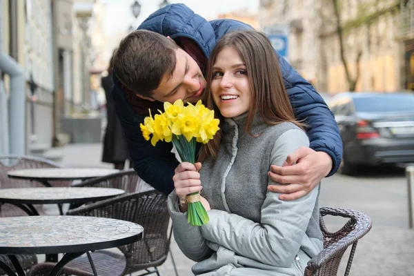 Couple in love in city. Young happy man and woman in spring city talking, walking, with bouquet of yellow flowers daffodils — Stock Photo, Image