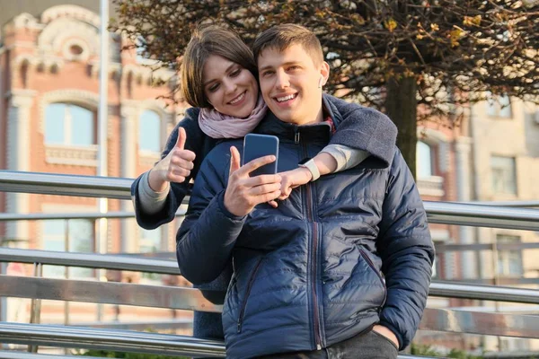 Retrato al aire libre de pareja mostrando signo de gesto ok. Joven hombre y mujer con teléfono inteligente en auriculares, fondo de la ciudad hora dorada —  Fotos de Stock