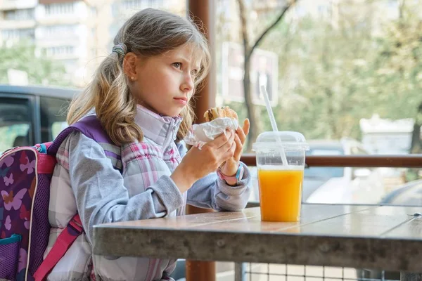 Ao ar livre retrato bonita estudante 8, 9 anos com cheeseburger e suco de laranja, fast food restaurante fundo — Fotografia de Stock