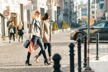 Kentsel tarzı, iki genç gülümseyen moda kadın alışveriş çantaları, güneşli sonbahar günü, altın saat ile bir şehir sokak boyunca yürüyüş