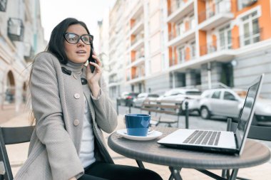 Güzel bir kız, gözlüklü, kalın bir palto takıyor, dışarıda bir kafede oturuyor, bir bardak sıcak içecekle dizüstü bilgisayarında oturuyor, cep telefonuyla konuşuyor, şehir arka planında.