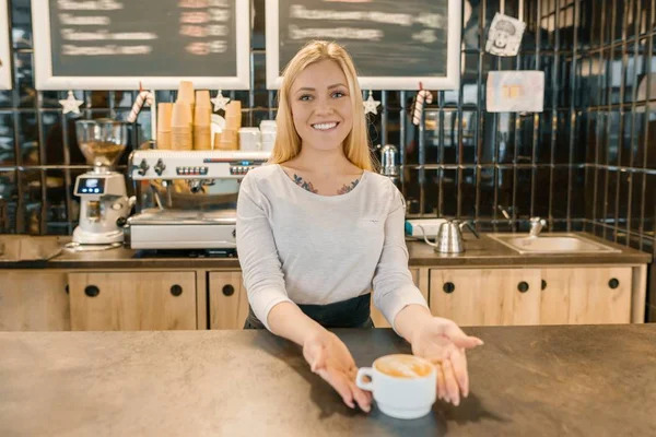 若い笑顔のバリスタの女の子が淹れたてのコーヒーを飲みながら。エプロンを着たブロンドの女性コーヒーショップのバーカウンターの近く — ストック写真