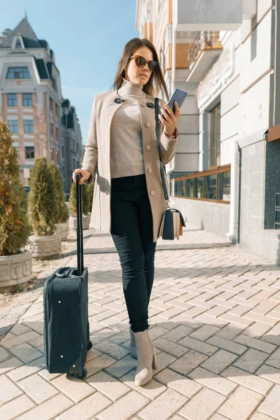 Portrait of traveling young woman with mobile phone and suitcase, fashionable girl on the city street, wearing warm coat, sunny autumn day